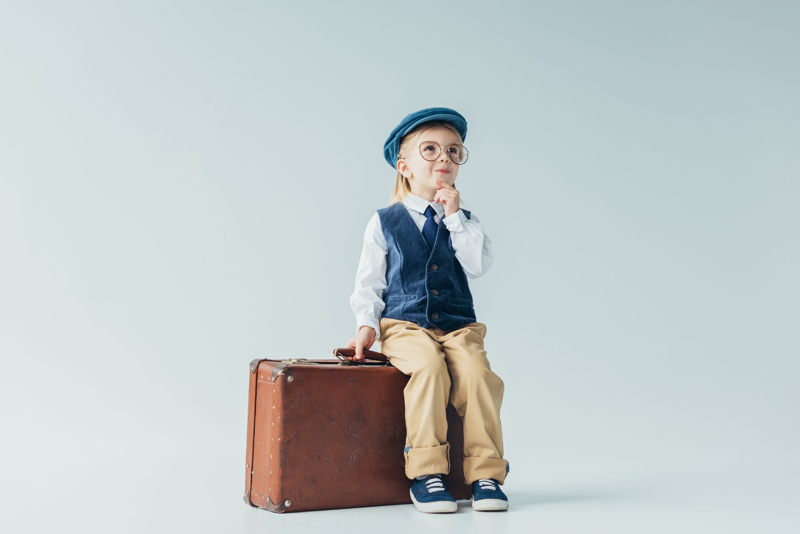 pensive kid in retro vest and cap sitting on suitcase on grey background